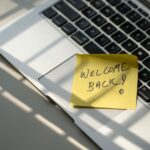 Ordenador portátil encendido con un post-it amarillo pegado que dice "Welcome back", sobre un escritorio minimalista.