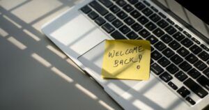 Ordenador portátil encendido con un post-it amarillo pegado que dice "Welcome back", sobre un escritorio minimalista.