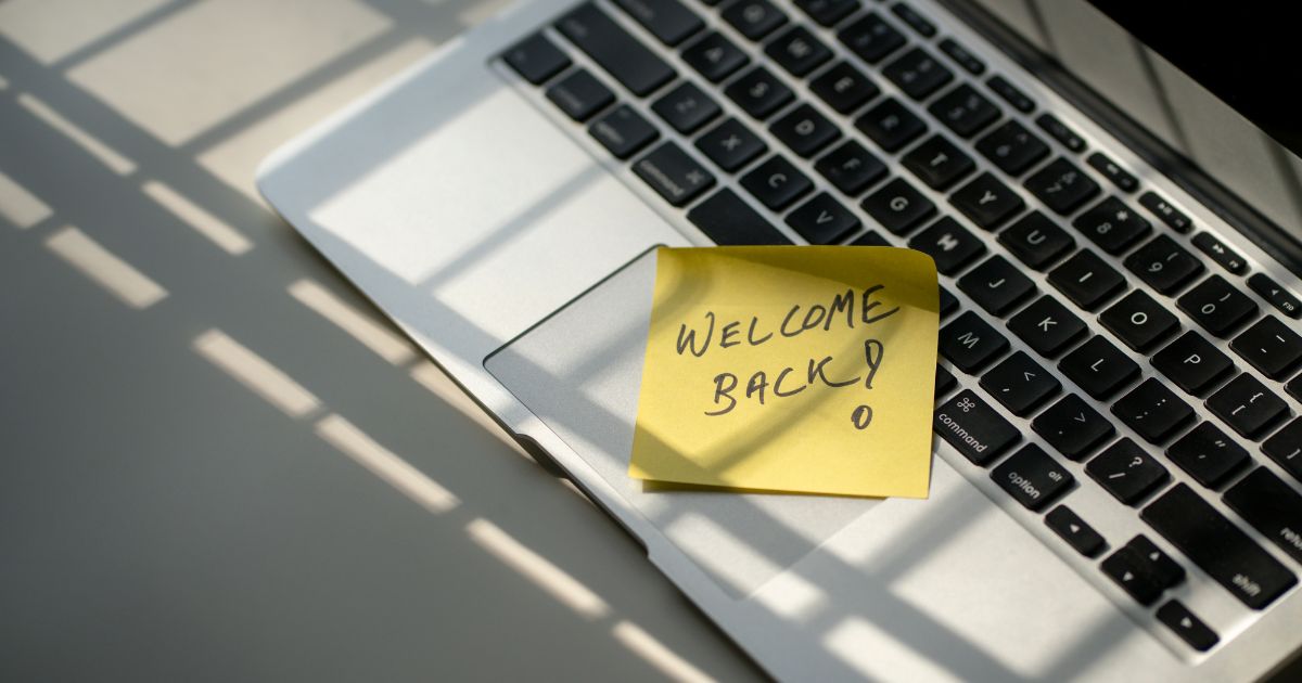 Ordenador portátil encendido con un post-it amarillo pegado que dice "Welcome back", sobre un escritorio minimalista.